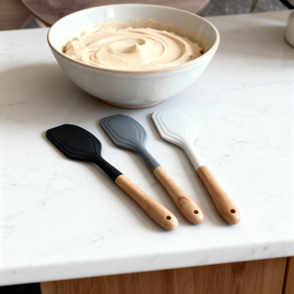 Trendy Shop silicone scraper spatulas in black, grey, and white with wooden handles arranged on a marble countertop.
